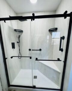 Walk-in shower with a black framed glass sliding door, light large-format wall panels, grab bars and a built-in corner bench