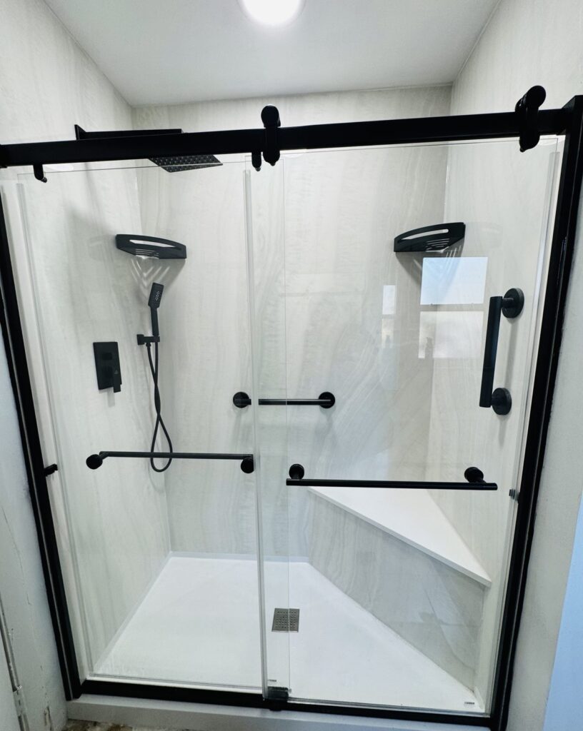 Walk-in shower with a black framed glass sliding door, light large-format wall panels, grab bars and a built-in corner bench
